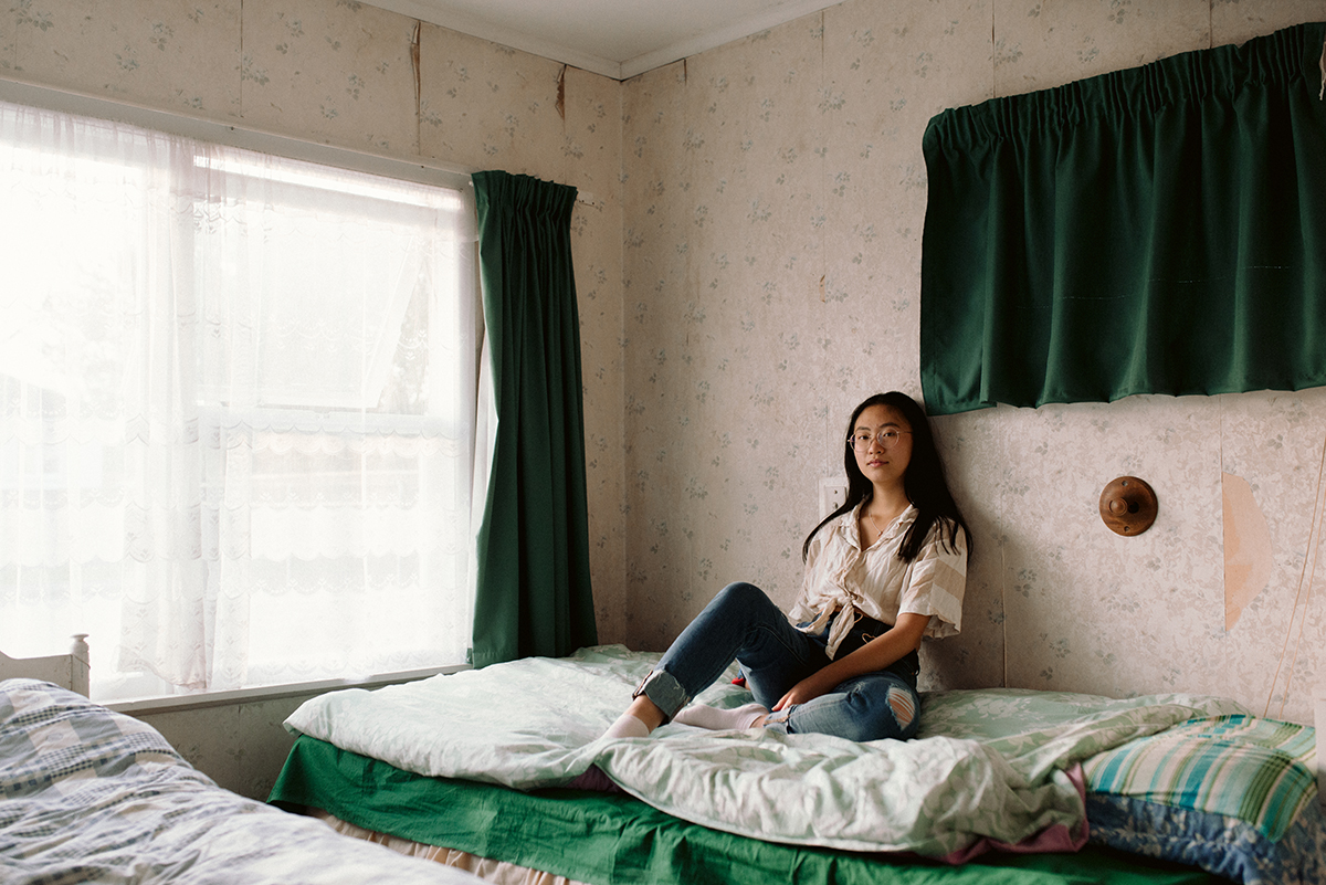 Aimee in jeans and buttoned shirt, sitting on her bed, leaning against the wall with patterned wallpaper
