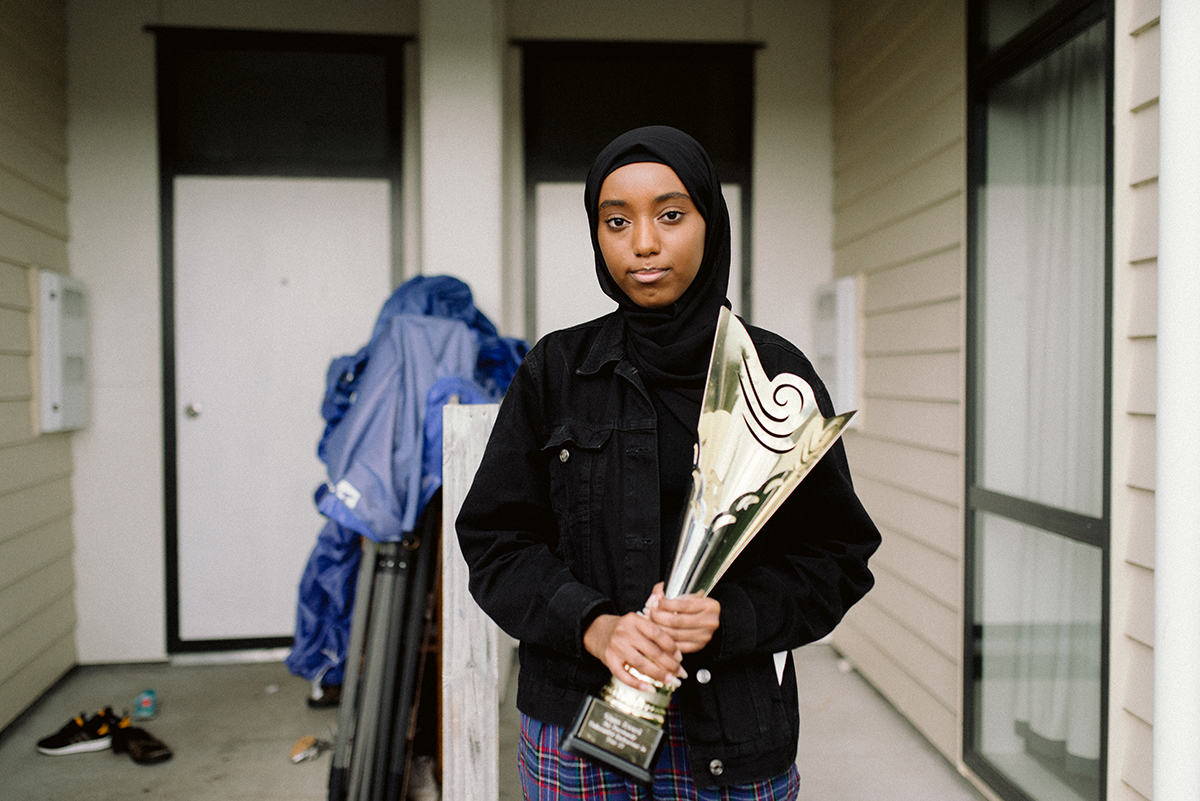 Tasnim in a black hijab, holding a trophy and standing in front of her front door
