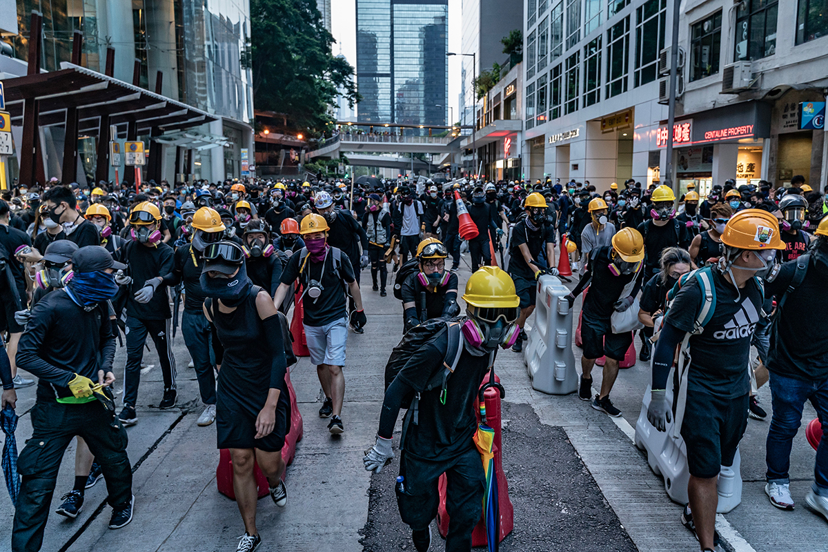 People in black clothing and yellow safety helmets walk down street dragging supplies