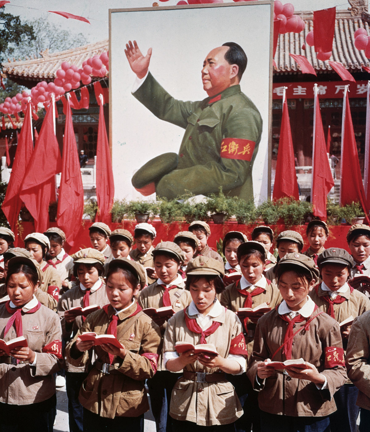 Children in uniform standing in rows, holding red booklets in front of painting of Mao Zedong